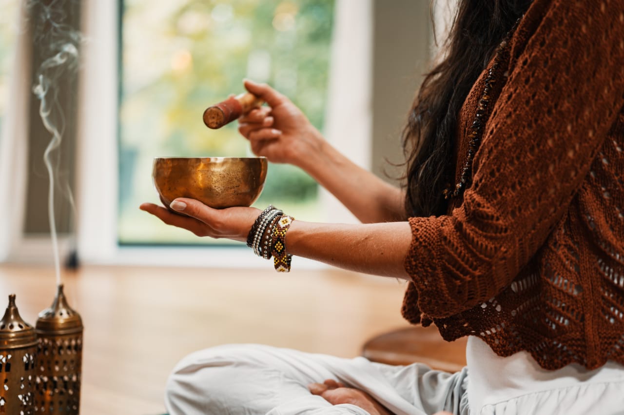 An image of a woman holding a brass bowl in her left hand and a wooden stir-stick in her right, while burning incense and sitting cross-legged on the floor